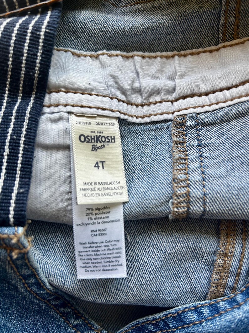 May include: Close-up of a pair of denim overalls with a white and navy blue striped strap. The tag reads "OSHKOSH B'gosh" and "4T". The overalls are made in Bangladesh and are composed of cotton, polyester, and elastane.