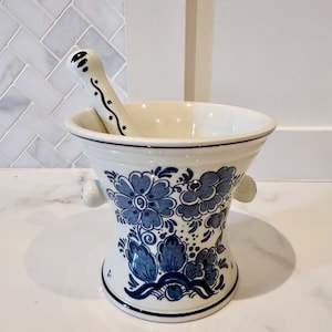 Może przedstawiać: Biały ceramiczny zestaw moździerza i tłuczka. Moździerz ma niebieskie kwiatowe wzory i małe uchwyty. Tłuczek jest biały z czarnymi detalami w kropki i linie. Zestaw przeznaczony jest do mielenia ziół i przypraw.