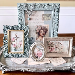 May include: A collection of ornate picture frames in various shapes and sizes, arranged on a decorative silver tray. The frames are in shades of blue and cream, with vintage-style artwork featuring roses, anchors, and a portrait. A silver necklace is draped over one frame.