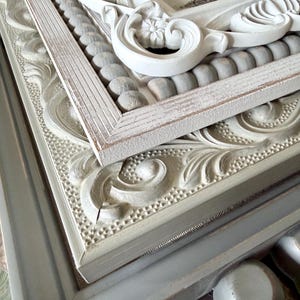 May include: A stack of ornate picture frames in various shades of white and gray. The frames feature detailed carvings, including floral and scroll designs, and some have beaded accents. The frames are stacked, showcasing their different textures and styles.