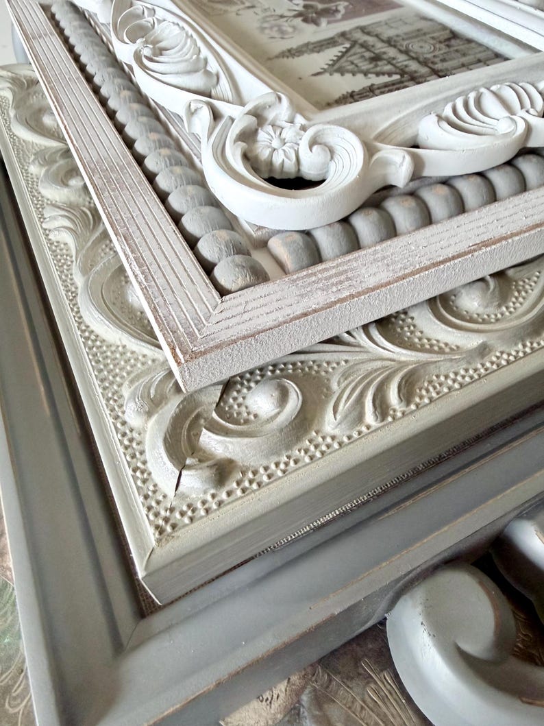May include: A stack of ornate picture frames in various shades of white and gray. The frames feature detailed carvings, including floral and scroll designs, and some have beaded accents. The frames are stacked, showcasing their different textures and styles.