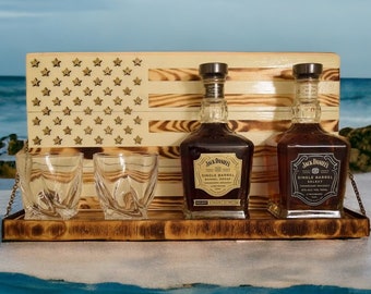 May include: A wooden tray with an American flag design features two bottles of Jack Daniel's Single Barrel Select Tennessee Whiskey. The tray also includes four clear glass shot glasses. The tray is sitting on a sandy beach with the ocean in the background.