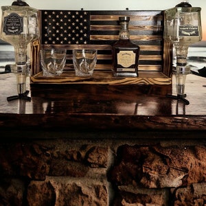 May include: A rustic wooden bar setup featuring a carved American flag backdrop. It includes two whiskey dispensers, two glasses, and a bottle of Jack Daniel's whiskey. The bar is set against a sunset backdrop, with a stone brick base.