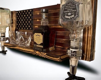 Puede incluir: Un dispensador de licor de pared de madera con un diseño de bandera estadounidense. El dispensador tiene dos botellas de vidrio transparente con etiquetas negras que dicen "Jack Daniel's Single Barrel Tennessee Whiskey Select" y "Jack Daniel's Tennessee Whiskey Single Barrel". El dispensador también tiene dos vasos de chupito de vidrio transparente.
