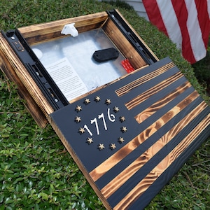 Puede incluir: Una bandera americana de madera con fondo negro y estrellas doradas. La bandera está abierta para revelar un compartimento oculto. El compartimento está forrado con un material negro y tiene una corredera de metal.