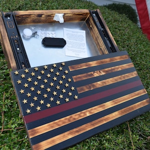 Puede incluir: Una bandera americana de madera con una franja roja en el medio, un compartimento oculto y un deslizador de metal negro. La bandera se muestra sobre un fondo verde.