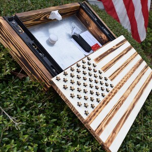 Puede incluir: Una bandera americana de madera con un compartimento oculto. La bandera está pintada de blanco con vetas de madera marrón que se ven a través. Las estrellas son negras y las rayas son blancas. El compartimento está abierto y revela una caja de metal.
