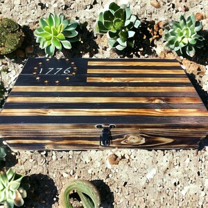 Puede incluir: Una caja de madera con diseño de bandera. La caja es rectangular con tapa con bisagras y cierre negro. El diseño de la bandera presenta rayas negras y beige, con el año "1776" y estrellas en la esquina superior izquierda. La caja está rodeada de suculentas.