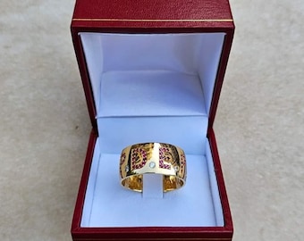 Puede incluir: Un anillo de oro con una banda de piedras preciosas rojas y una sola piedra blanca en el centro. El anillo está en una caja de terciopelo rojo.