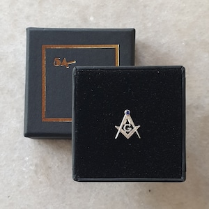 May include: A silver Masonic pin with a blue gem, displayed in a black velvet-lined box. The pin features a square and compass design with the letter "G" in the center. The box is next to a black box with gold trim and the text "5A".