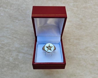 May include: A silver signet ring with a white enamel top featuring a gold five-pointed star in a circle. The ring is in a red velvet box.