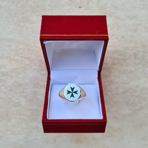 Op de afbeelding: Een zilveren ring met een witte ovale steen met een groen kruis. De ring zit in een rode fluwelen ringdoos.
