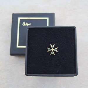 Puede incluir: Un pequeño pin de color dorado con un diseño de cruz de Malta de esmalte verde. El pin está en una caja de terciopelo negro con borde dorado.