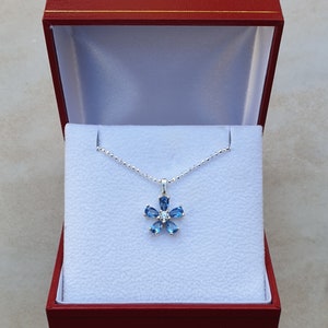 May include: A silver necklace with a blue topaz flower pendant. The pendant is set with six blue topaz stones and one clear stone in the center. The necklace is in a red jewelry box.