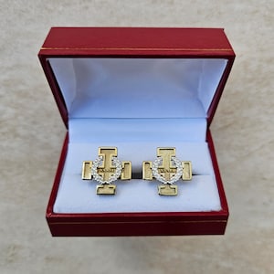 May include: A pair of gold cufflinks in a red box. The cufflinks feature a gold cross with a silver wreath and the word "XXVII" in the center.