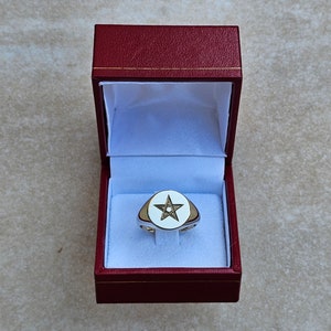 Puede incluir: Un anillo de oro con una piedra ovalada blanca con un diseño de estrella de cinco puntas, presentado en una caja de anillo de terciopelo rojo.