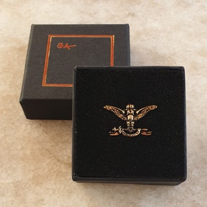 Può includere: Uno spillo dorato con una creatura alata e uno stendardo con la scritta "5A". Lo spillo è esposto in una scatola di velluto nero con una scatola esterna nera.