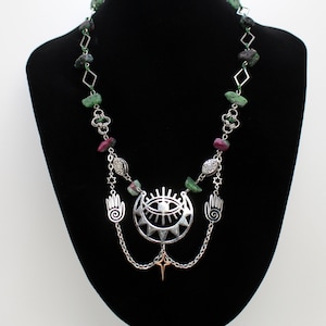 May include: A silver necklace with a green and pink gemstone pendant featuring a stylized eye, two hands, and a star. The necklace is on a black velvet background.