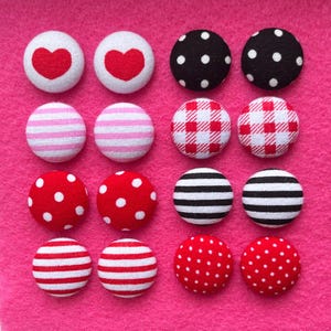 Puede incluir: Un conjunto de 12 botones cubiertos de tela en varios patrones. Los botones son rojos, blancos, negros y rosas con lunares, rayas, cuadros y corazones.