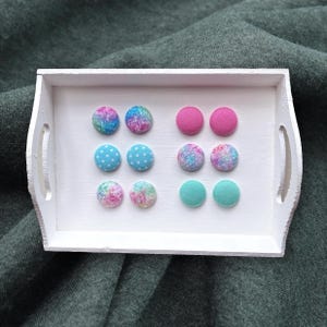 May include: A white wooden tray holds an assortment of colorful, round fabric-covered buttons. The buttons feature various designs, including polka dots, glitter, and solid colors like pink and teal. The tray sits on a dark green fabric surface.