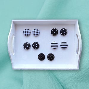 May include: A white tray displays six pairs of black and white patterned stud earrings. Patterns include gingham, polka dots, stars, and stripes. The earrings are arranged on a white tray with a light green background.
