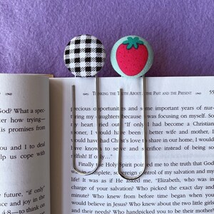 May include: Two silver bookmark clips with fabric covers. One clip has a black and white checkered pattern, the other has a red strawberry on a green background. The text "TINKING THE TUTH ABOUT THE PAST AND THE PRESENT" is visible in the background.