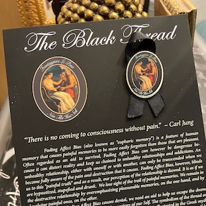 Puede incluir: Una imagen en blanco y negro de un pin con una cinta negra. El pin presenta una ilustración en blanco y negro de una mujer dando un hilo a un hombre. El texto en el pin dice "Fronteggiare Le Pena (Confront the Pain)". El texto en la tarjeta dice "The Black Thread" e incluye una cita de Carl Jung: "No hay conciencia sin dolor."