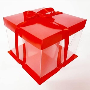 May include: A red gift box with a clear plastic window and a red ribbon tied in a bow. The box is perfect for presenting a cake or other baked goods.