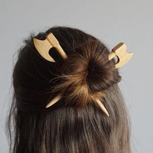 May include: A pair of wooden hair sticks with axe-shaped tops. The sticks are crossed and used to hold a bun of brown hair.