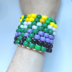 Könnte beinhalten: Ein mehrfarbiges Perlenarmband mit gelben, grünen, weißen, lila, schwarzen und grünen Perlen. Das Armband besteht aus kleinen, runden Perlen, die auf einer weißen Schnur aufgefädelt sind.