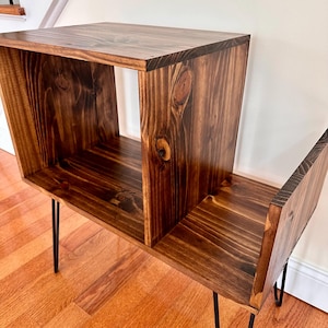 Op de afbeelding: Een donker gebeitste houten bijzettafel met een vak en een onderste plank. De tafel heeft zwarte haarspeldpoten en is gemaakt van massief hout. De houtnerf is zichtbaar.