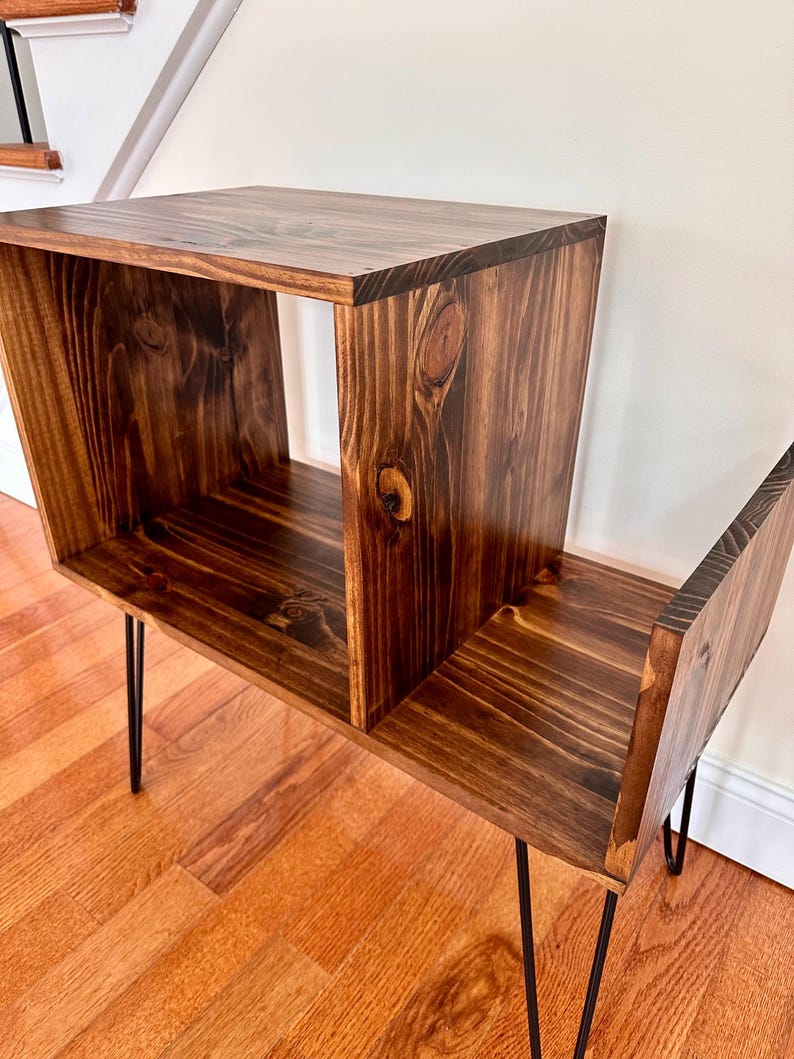 Op de afbeelding: Een donker gebeitste houten bijzettafel met een vak en een onderste plank. De tafel heeft zwarte haarspeldpoten en is gemaakt van massief hout. De houtnerf is zichtbaar.