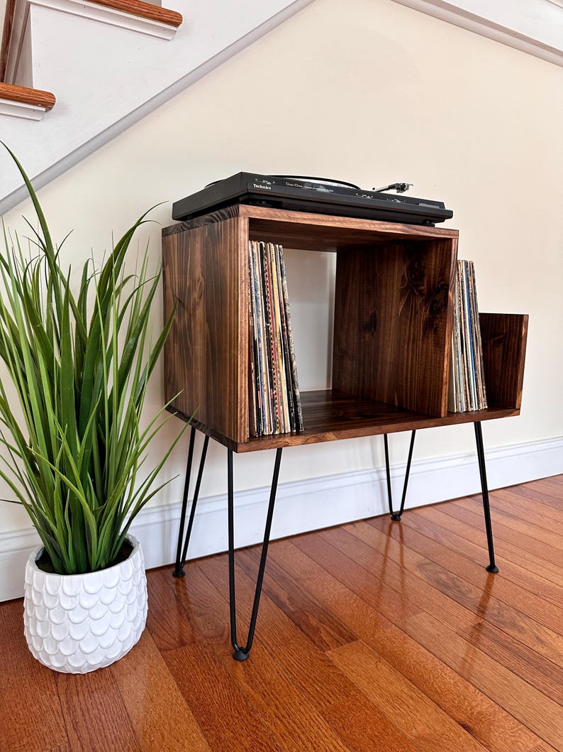 Op de afbeelding: Een donker gebeitste houten platenspelerstandaard met zwarte haarspeldpoten. De standaard heeft een vierkante bovenkant met een platenspeler en opslag voor vinylplaten. Links staat een witte potplant.