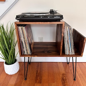 Op de afbeelding: Een donker gebeitste houten platenspelerstandaard met zwarte haarspeldpoten. De standaard heeft twee compartimenten voor vinylplaten en een bovenkant voor een zwarte platenspeler. Een potplant staat links.