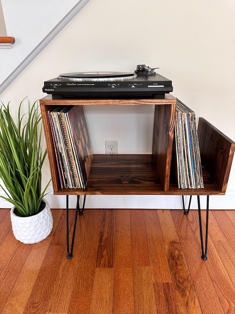 Op de afbeelding: Een donker gebeitste houten platenspelerstandaard met zwarte haarspeldpoten. De standaard heeft twee compartimenten voor vinylplaten en een bovenkant voor een zwarte platenspeler. Een potplant staat links.
