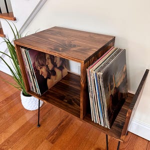 Op de afbeelding: Een donker gebeitste houten platenopslag met zwarte haarspeldpoten. De unit heeft twee compartimenten, elk met vinylplaten. De unit staat op een hardhouten vloer, met een trap en een plant op de achtergrond.