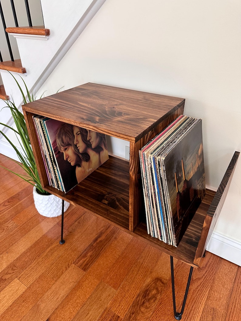 Op de afbeelding: Een donker gebeitste houten platenopslag met zwarte haarspeldpoten. De unit heeft twee compartimenten, elk met vinylplaten. De unit staat op een hardhouten vloer, met een trap en een plant op de achtergrond.