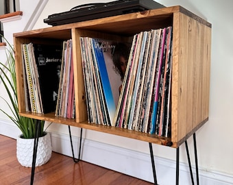 Puede incluir: Un mueble de madera para tocadiscos con patas de horquilla negras. El mueble tiene dos compartimentos llenos de discos de vinilo y un tocadiscos negro en la parte superior. La madera tiene un acabado natural.