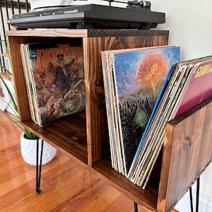 Op de afbeelding: Een donker gebeitste houten platenspelerstandaard met zwarte haarspeldpoten. De standaard heeft compartimenten voor vinylplaten en een zwarte platenspeler bovenop. De platen zijn zichtbaar en tonen albumhoezen.