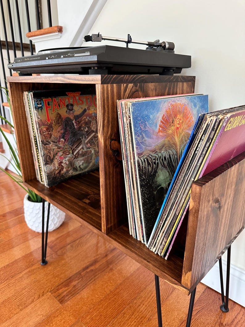 Op de afbeelding: Een donker gebeitste houten platenspelerstandaard met zwarte haarspeldpoten. De standaard heeft compartimenten voor vinylplaten en een zwarte platenspeler bovenop. De platen zijn zichtbaar en tonen albumhoezen.