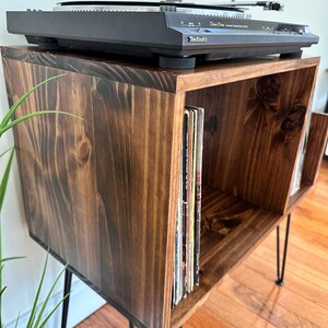 Op de afbeelding: Een donkerbruine houten platenspelerstandaard met zwarte haarspeldpoten. Bovenop staat een zwarte Technics platenspeler. Vinylplaten worden opgeborgen in de open vakken eronder. De standaard is gemaakt van gebeitst hout.