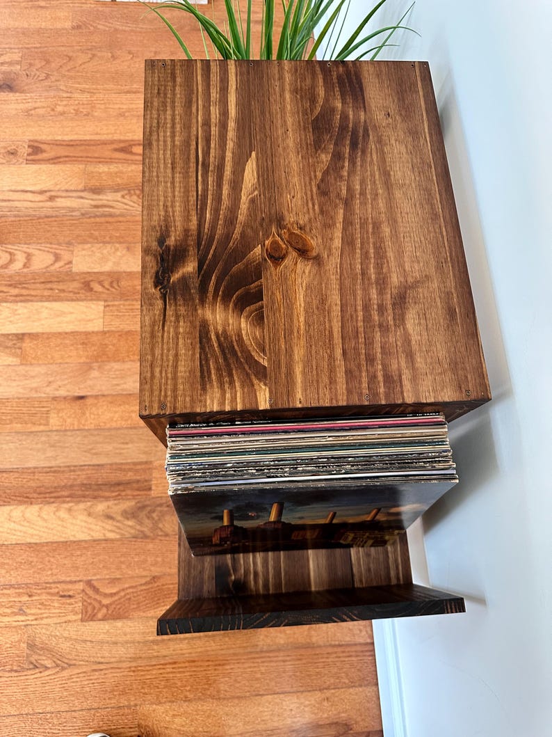 Op de afbeelding: Een donker gebeitste houten platenopslag met een ingebouwde plantenbak. De unit heeft een bovenste gedeelte voor planten en een onderste plank voor vinylplaten. De houtnerf is zichtbaar.