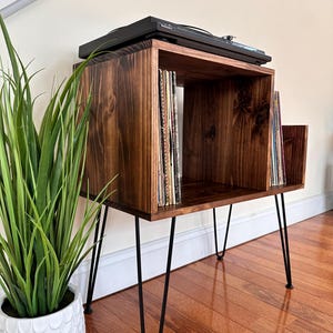 Op de afbeelding: Een donker gebeitste houten platenspelerstandaard met zwarte haarspeldpoten. De standaard heeft een vierkante bovenkant met een vak voor platen en een plank voor een platenspeler. Bovenop staat een zwarte platenspeler.