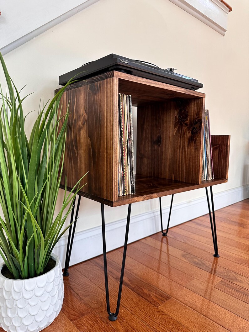 Op de afbeelding: Een donker gebeitste houten platenspelerstandaard met zwarte haarspeldpoten. De standaard heeft een vierkante bovenkant met een vak voor platen en een plank voor een platenspeler. Bovenop staat een zwarte platenspeler.