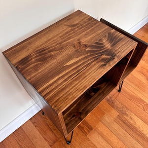 Op de afbeelding: Een gebeitste houten bijzettafel met een donkerbruine afwerking. De tafel heeft een rechthoekige vorm met een open plank en staat op zwarte haarspeldpoten. De houtnerf is zichtbaar, wat textuur aan het stuk toevoegt. De tafel is ontworpen voor opslag en display. Afmetingen: 60cm x 40cm x 40cm.