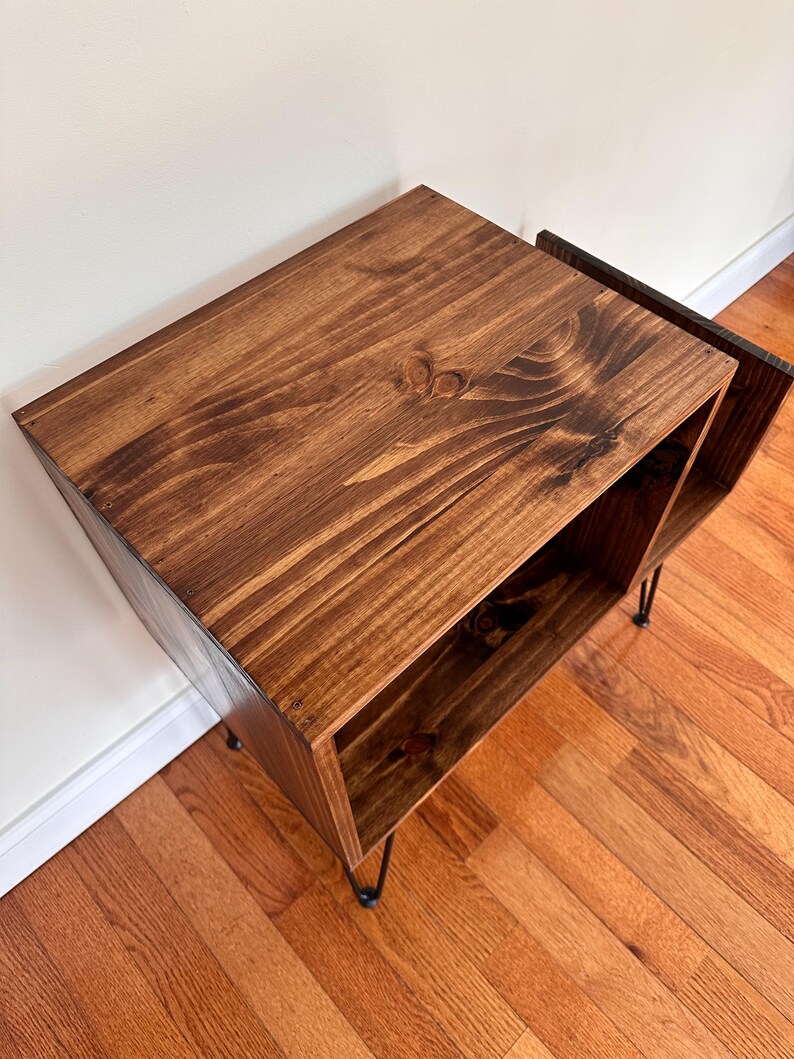 Op de afbeelding: Een gebeitste houten bijzettafel met een donkerbruine afwerking. De tafel heeft een rechthoekige vorm met een open plank en staat op zwarte haarspeldpoten. De houtnerf is zichtbaar, wat textuur aan het stuk toevoegt. De tafel is ontworpen voor opslag en display. Afmetingen: 60cm x 40cm x 40cm.