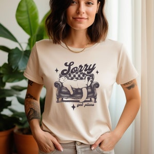 May include: A beige t-shirt with a graphic of a cat lounging on a couch. The text "Sorry got plans" is printed below the cat.