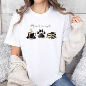 May include: White t-shirt with the phrase "My needs are simple." featuring a coffee cup, paw print, and books. The coffee cup is black with steam rising. The paw print is black and brown. Two books are stacked.