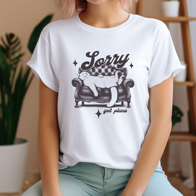 May include: White t-shirt with a graphic of a cat sleeping on a couch. The text "Sorry got plans" is printed below the cat.