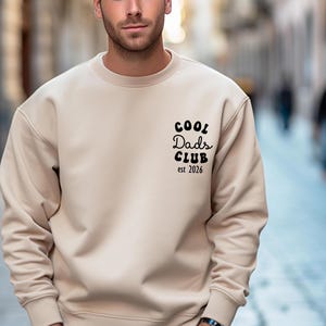 May include: A beige crew neck sweatshirt with the text "COOL Dads CLUB est 2026" printed in black on the upper left chest. The sweatshirt is a solid color and has long sleeves.